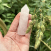 A hand holds a polished Rose Quartz tower in soft pink tones, surrounded by lush green grass and delicate flowering stalks, symbolizing love, peace and nature's embrace.