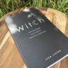 Black book titled "Witch" with a crescent moon replacing the "c," lying on a wooden tray surrounded by grass.