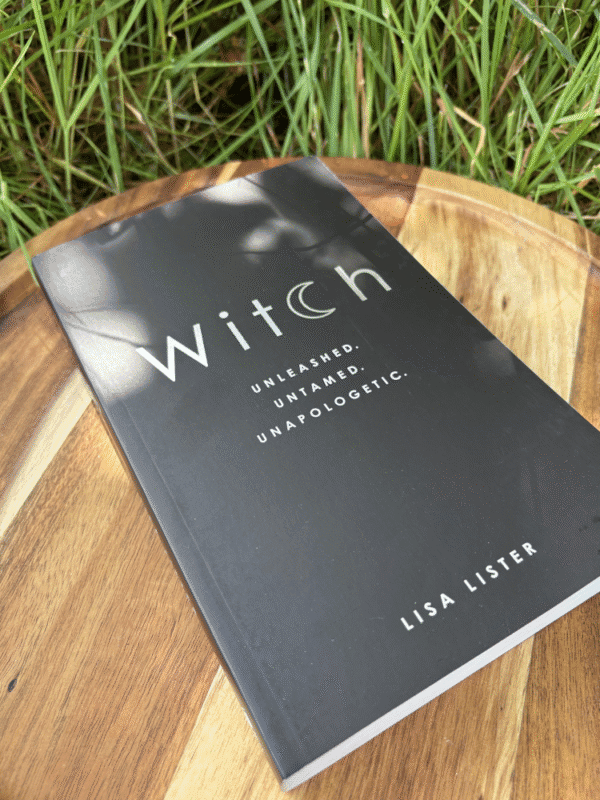 Black book titled "Witch" with a crescent moon replacing the "c," lying on a wooden tray surrounded by grass.