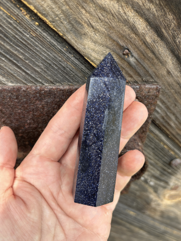 A hand holding a shimmering blue sandstone crystal tower, sparkling like a midnight sky against rustic wooden background.