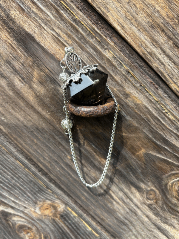 Obsidian-colored resin crystal pendulum with Tree of Life charm, resting on rustic wood and metal.
