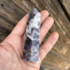 A hand holds a polished Chevron Amethyst tower, showcasing stunning violet and white zigzag patterns. The background of rustic wood enhances the stone's spiritual and intuitive energy.