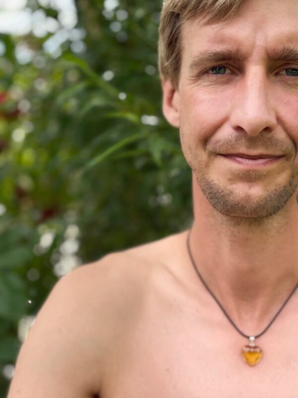Person wearing a golden heart pendant on a black waxed cord necklace, standing shirtless outdoors with soft green foliage in the background