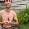 A shirtless person sits peacefully in meditation outdoors, eyes closed, holding a black obsidian tower at the heart center. Dressed in vibrant rainbow-patterned pants, surrounded by flowers and greenery, embodying grounding, balance, and inner presence.