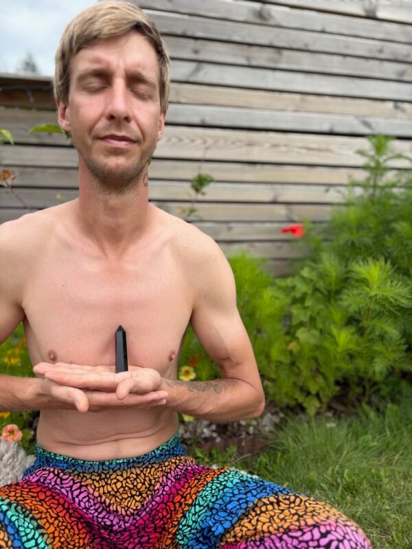 A shirtless person sits peacefully in meditation outdoors, eyes closed, holding a black obsidian tower at the heart center. Dressed in vibrant rainbow-patterned pants, surrounded by flowers and greenery, embodying grounding, balance, and inner presence.