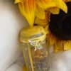 Glass jar filled with herbs and flowers, sealed with golden beeswax and adorned with a dragonfly charm, surrounded by vibrant sunflowers – a spell jar radiating joy, positivity, and solar magic.