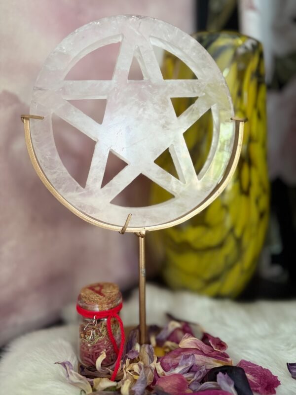 White quartz pentacle standing in golden frame with red intention jar and dried petals around.