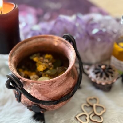 Witchy scene with a small copper cauldron filled with herbs, placed on faux fur next to a candle and amethyst crystals.