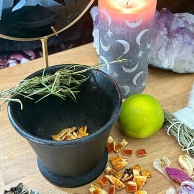 Antique cast iron cauldron on an altar with herbs, dried citrus, and a lit moon-phase candle, surrounded by crystals and petals.