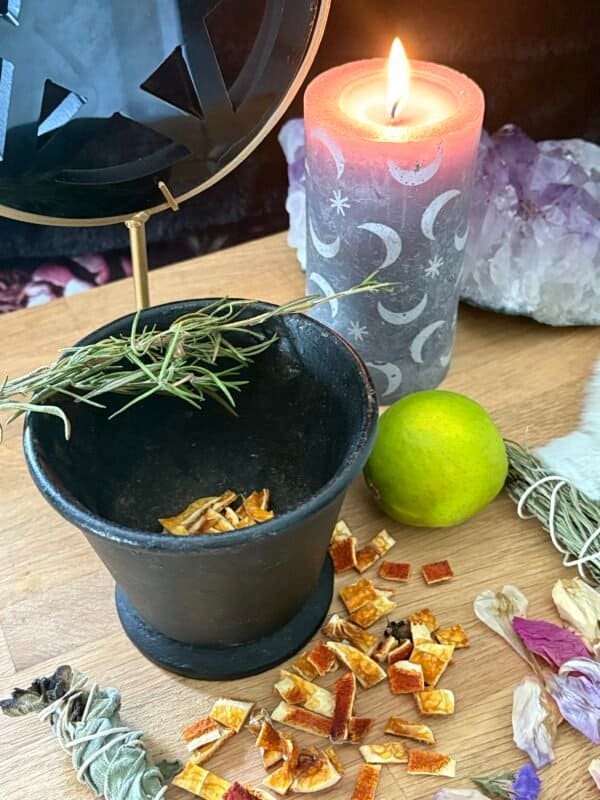 Antique cast iron cauldron on an altar with herbs, dried citrus, and a lit moon-phase candle, surrounded by crystals and petals.
