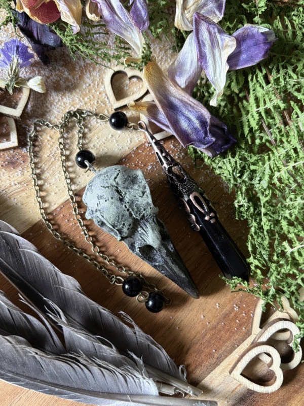Obsidian crystal pendulum with raven skull detail, photographed against natural background, gothic witch style