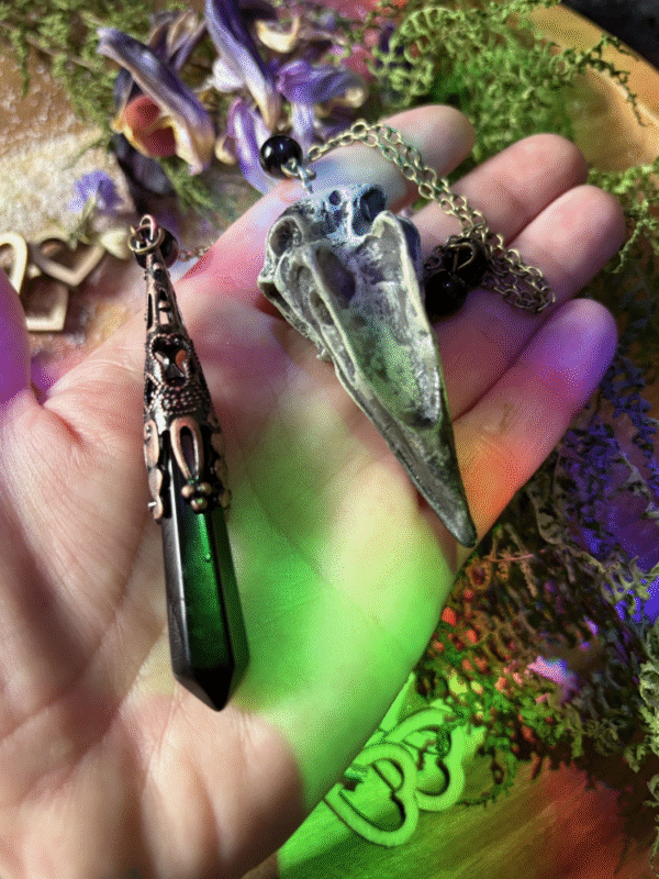 Obsidian crystal pendulum with raven skull detail, photographed against natural background, gothic witch style.
