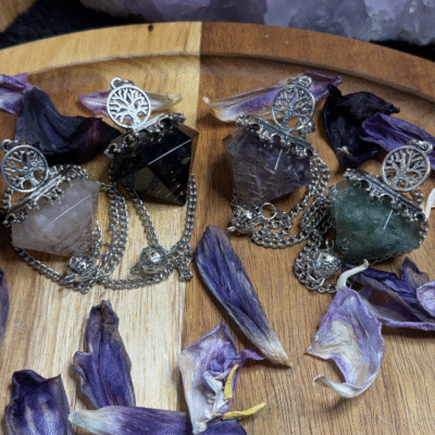 Four mystical resin pendulums with crystal chips, copper spirals, and silver Tree of Life charms, surrounded by dried violet petals on a wooden surface with amethyst crystals in the background.