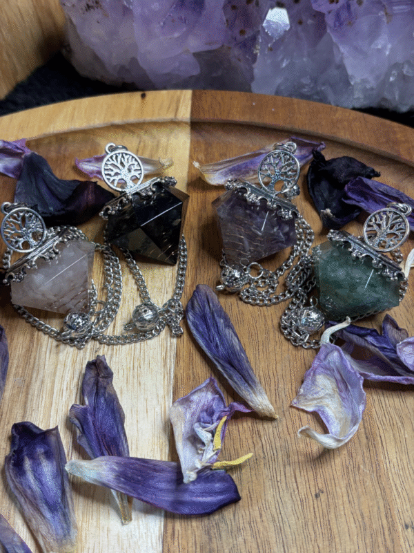 Four mystical resin pendulums with crystal chips, copper spirals, and silver Tree of Life charms, surrounded by dried violet petals on a wooden surface with amethyst crystals in the background.