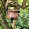 A vintage copper cauldron with black iron handle and legs, hanging from a tree branch surrounded by green leaves – perfect for witchy rituals and nature altars.