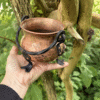 Close-up of a hand holding a small vintage copper cauldron with iron legs and handle, framed by trees and greenery – an ideal tool for mystical work and rustic decor.