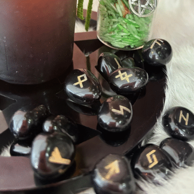 Black obsidian rune stones with golden symbols displayed in a magical setting beside a candle and spell jar.