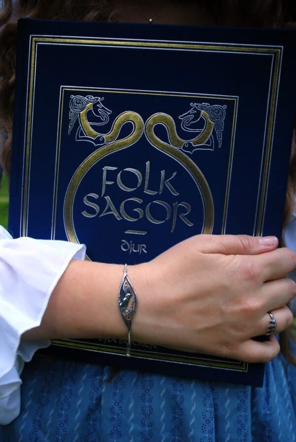 Nattuggla" silver bracelet with owl and moon, styled with Nordic-inspired "Folksagor" book in blue and gold.