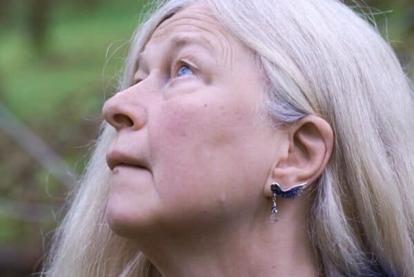 Older woman wearing silver owl earrings, inspired by Nordic mythology, looking upwards in nature.