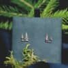 Close-up of silver pine tree stud earrings displayed on a deep green velvet background with forest elements.