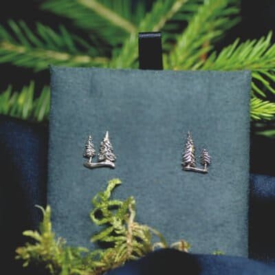 Close-up of silver pine tree stud earrings displayed on a deep green velvet background with forest elements.