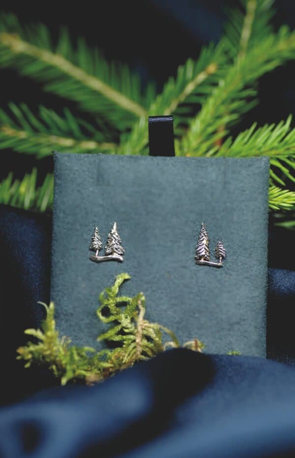 Close-up of silver pine tree stud earrings displayed on a deep green velvet background with forest elements.