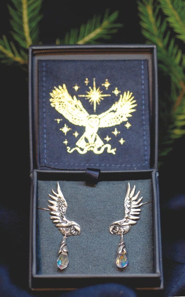 Silver owl earrings with opal glass drops displayed in an elegant jewelry box with golden owl emblem.